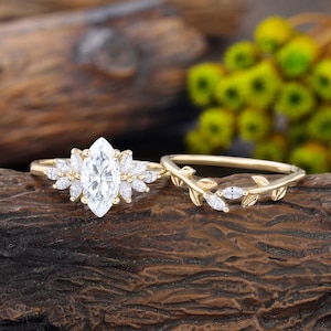 May include: A gold ring set featuring a central marquise-cut stone surrounded by smaller marquise-cut stones in a cluster arrangement, paired with a matching gold band designed with a delicate leaf motif and small inlaid stones.