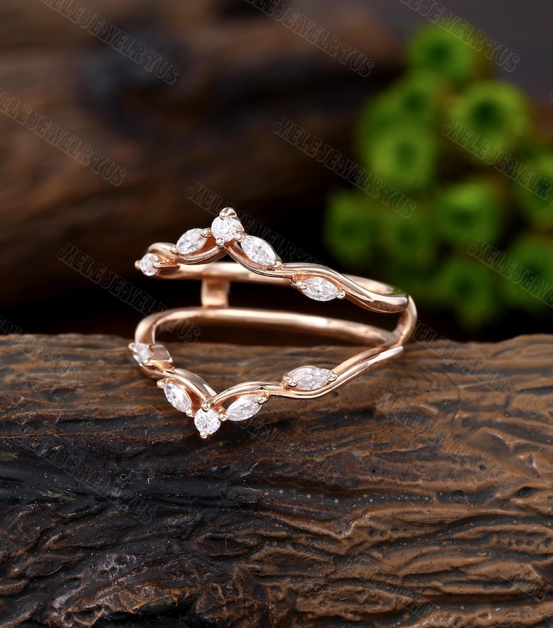 May include: Rose gold ring with a delicate marquise diamond design. The ring is a split shank style, with two bands that meet at the top to create a V-shape. The diamonds are set in a pave setting, and the ring has a polished finish.