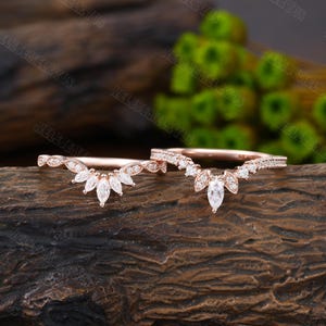 May include: Two delicate rose gold wedding bands with a marquise-shaped center stone and smaller round stones set in a curved, v-shaped design.