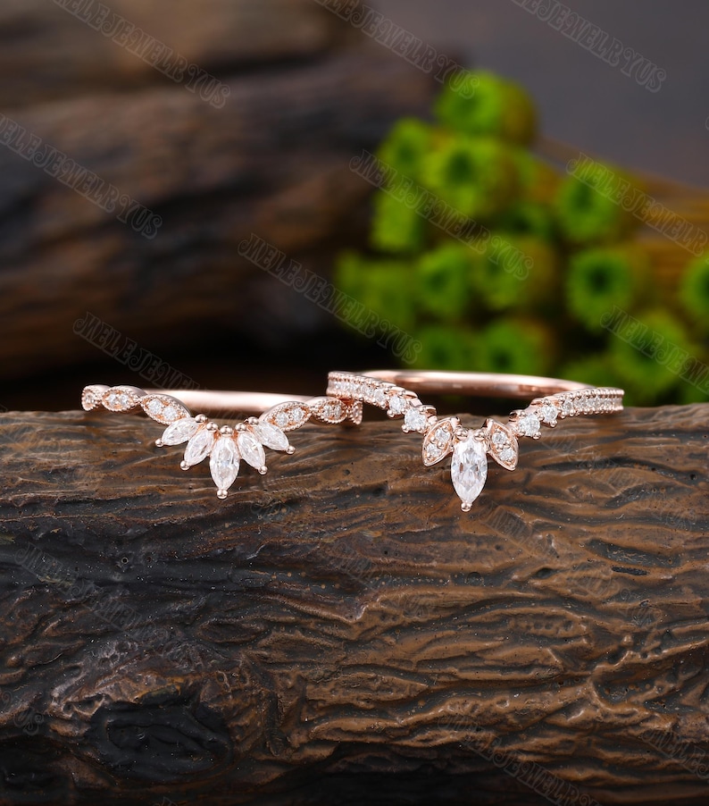 May include: Two delicate rose gold wedding bands with a marquise-shaped center stone and smaller round stones set in a curved, v-shaped design.