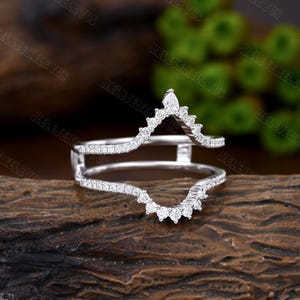 May include: A delicate, silver ring enhancer with a V-shaped design. The enhancer is adorned with small, sparkling diamonds, creating a shimmering effect. The ring is set against a rustic wooden background.