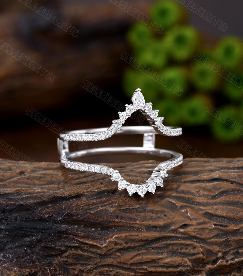 May include: A delicate, silver ring enhancer with a V-shaped design. The enhancer is adorned with small, sparkling diamonds, creating a shimmering effect. The ring is set against a rustic wooden background.