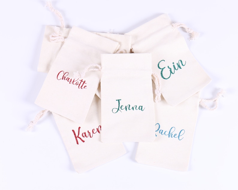 May include: Five small white fabric drawstring bags with different names embroidered on them in various colors. The names are Charlotte, Jenna, Erin, Rachel, and Karen.