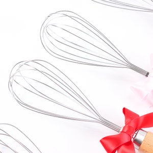 May include: Three silver wire whisks with wooden handles. The whisks are tied with red and pink ribbons.