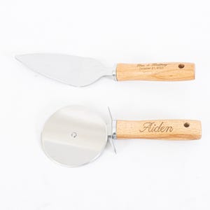 May include: A set of two kitchen tools with wooden handles. The larger tool is a cake server with a serrated edge. The smaller tool is a pizza cutter with a round blade. The handles are engraved with the text "Ben & Brittany October 21, 2023" and "Aiden".