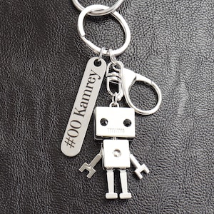 Peut inclure: Un porte-clés argenté en forme de robot avec un corps rectangulaire, des bras et des jambes. Le robot a des yeux noirs et une rangée de lignes pour la bouche. Une étiquette argentée avec le texte "#00 Kamrey" est suspendue au porte-clés.
