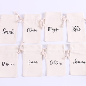 May include: Eight small white cotton drawstring bags with black lettering. The bags are arranged in two rows of four. The names on the bags are Sarah, Olivia, Maggie, Kiki, Rebecca, Lemon, Colleen, and Jenna.