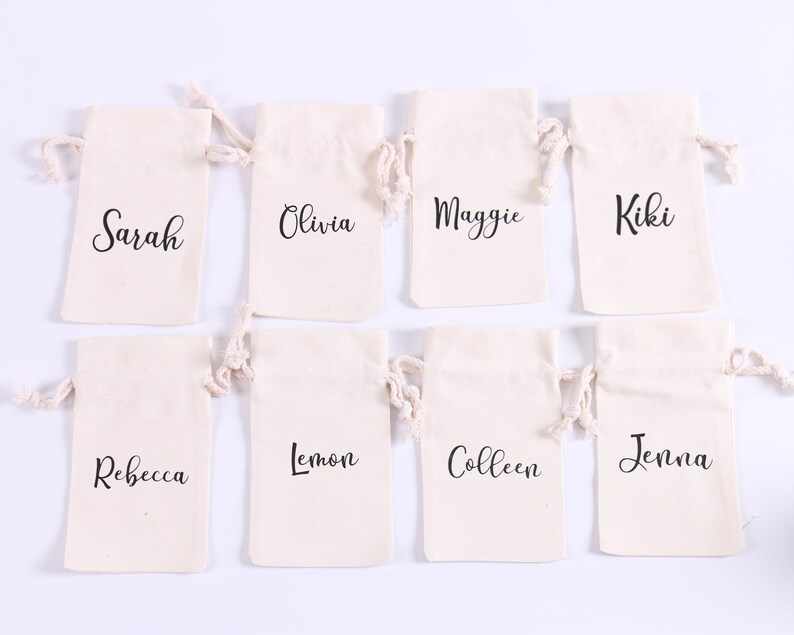 May include: Eight small white cotton drawstring bags with black lettering. The bags are arranged in two rows of four. The names on the bags are Sarah, Olivia, Maggie, Kiki, Rebecca, Lemon, Colleen, and Jenna.
