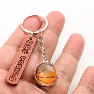May include: A basketball-themed keychain featuring a wooden tag with "Swissa #55" engraved in a bold font. The keychain also includes a round charm with a basketball design, suspended by a silver chain and ring.
