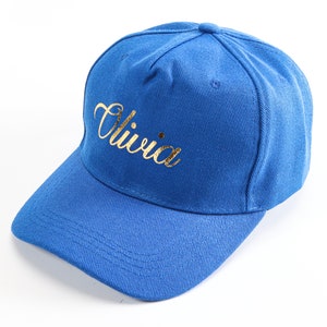 Può includere: Un berretto da baseball blu con il nome "Olivia" ricamato in oro sul davanti.