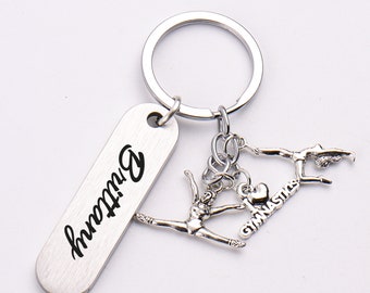 Op de afbeelding: Zilverkleurige sleutelhanger met een rechthoekig label met de naam "Brittany". De sleutelhanger heeft gymnastiekbedels en de woorden "I love Gymnastics".