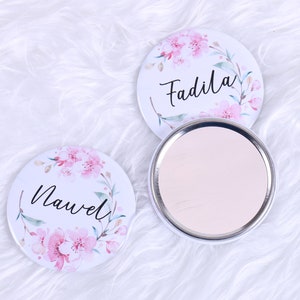 Op de afbeelding: Twee ronde compact spiegels met bloemenmotieven en de namen "Nawel" en "Fadila" erop gedrukt. De spiegels zijn wit met roze en groene bloemmotieven.