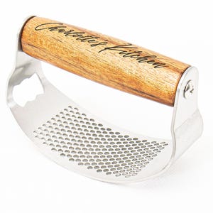 Pode incluir: Espremedor de alho em aço inoxidável com cabo de madeira. O cabo é gravado com as palavras "Charlottes Kitchen". O espremedor tem um design curvo com numerosos orifícios pequenos para espremer o alho.