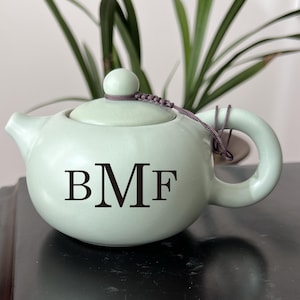 May include: A pale green ceramic teapot with a rounded body, a curved handle, and a spout. The lid is topped with a small knob and a decorative cord. The initials "BMF" are displayed in black on the front of the teapot.
