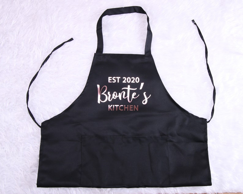 May include: Black kitchen apron with a large pocket and the text "EST 2020 Bronte's KITCHEN" in rose gold lettering.