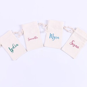 May include: Four small white fabric bags with drawstrings, each with a different name printed on the front in colorful lettering. The names are Lydia, Samantha, Myra, and Susan.