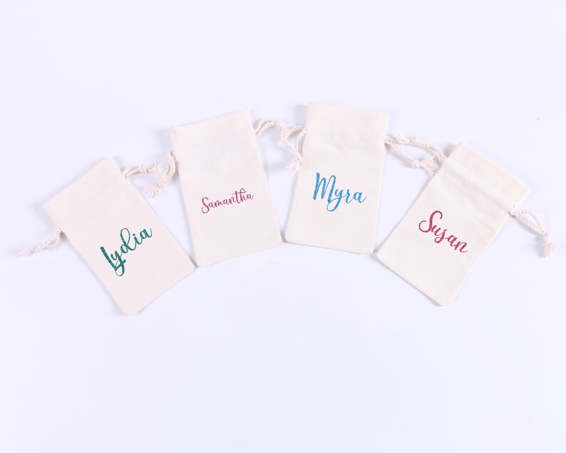 May include: Four small white fabric bags with drawstrings, each with a different name printed on the front in colorful lettering. The names are Lydia, Samantha, Myra, and Susan.