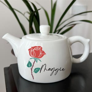 May include: White ceramic teapot with a red rose graphic and the name "Maggie" in black script. The teapot has a lid, spout, and handle. Green leaves and a brown stem accompany the rose design. A decorative item for tea lovers.