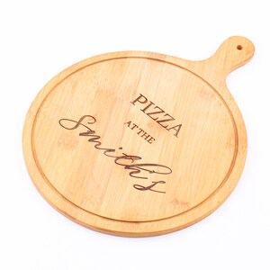 May include: A round, light brown bamboo pizza board with a handle. The board has a carved groove around the edge and features the engraved text "PIZZA at the Smiths".