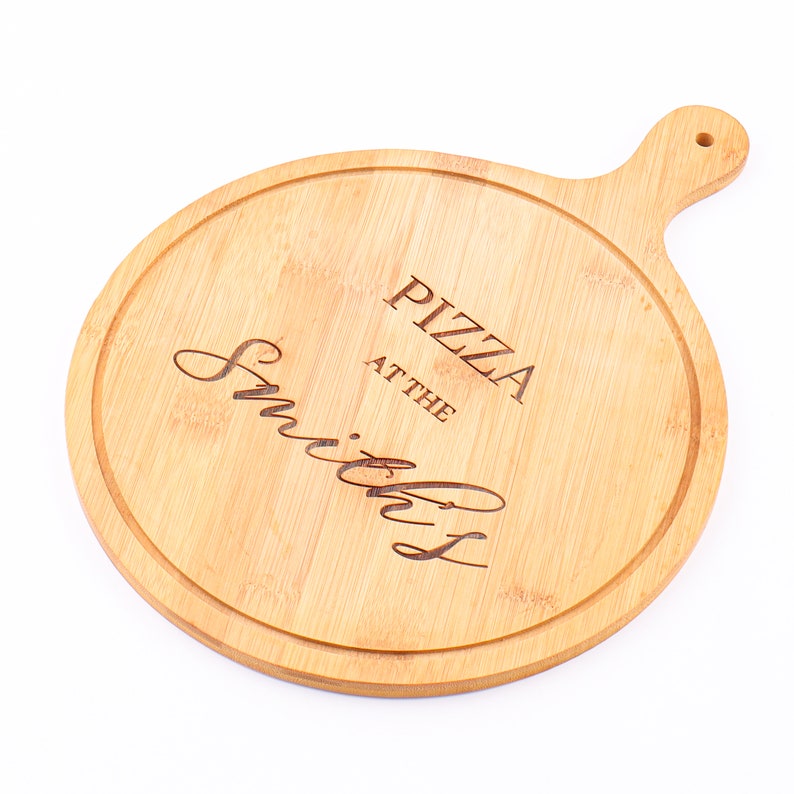 May include: A round, light brown bamboo pizza board with a handle. The board has a carved groove around the edge and features the engraved text "PIZZA at the Smiths".