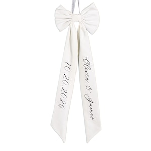 Op de afbeelding: Een witte satijnen strik met lange linten. De linten zijn bedrukt met de datum "10.20.2026" en de namen "Olivia & James" in zwart schrift. De strik is vastgebonden met een bijpassend wit lint.