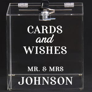 May include: Clear acrylic card box with a lock and key. The box has the words "CARDS and WISHES" and "MR. & MRS JOHNSON" in white lettering. The box is designed for weddings or special events.