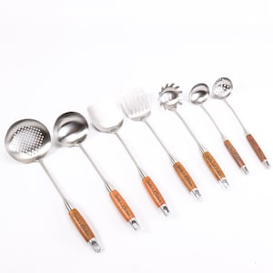 May include: Set of seven stainless steel kitchen utensils with wooden handles. The handles are engraved with "Julia's Kitchen". The utensils include a slotted spoon, a solid spoon, a spatula, a slotted turner, a pasta server, a ladle, and a skimmer.