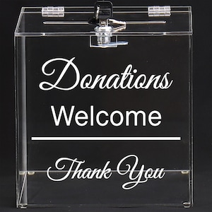 May include: Clear acrylic donation box with a lock and key. The box has the words "Donations Welcome Thank You" in white script. The box is rectangular and has a hinged lid.
