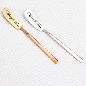 May include: Two butter knives with rounded blades and long handles. One is gold-coloured, the other silver. Both knives have the text "Tiffany & Tom" engraved on the blade.