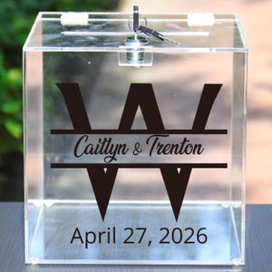 May include: Transparent acrylic box with a slot and a lock with keys. The front displays a black monogram, the names "Caitlyn & Trenton", and the date "April 27, 2026".