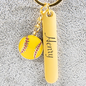 May include: A gold-coloured keyring featuring a softball charm and a rectangular tag. The softball charm is yellow with red stitching. The tag is engraved with the name "Henry" in black script.