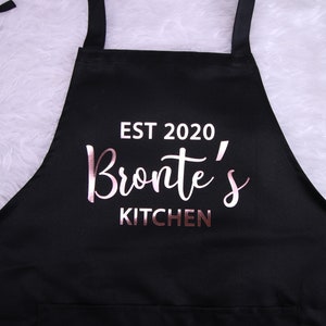 May include: Black apron with a white pocket and the text "EST 2020 Bronte's KITCHEN" in rose gold lettering.
