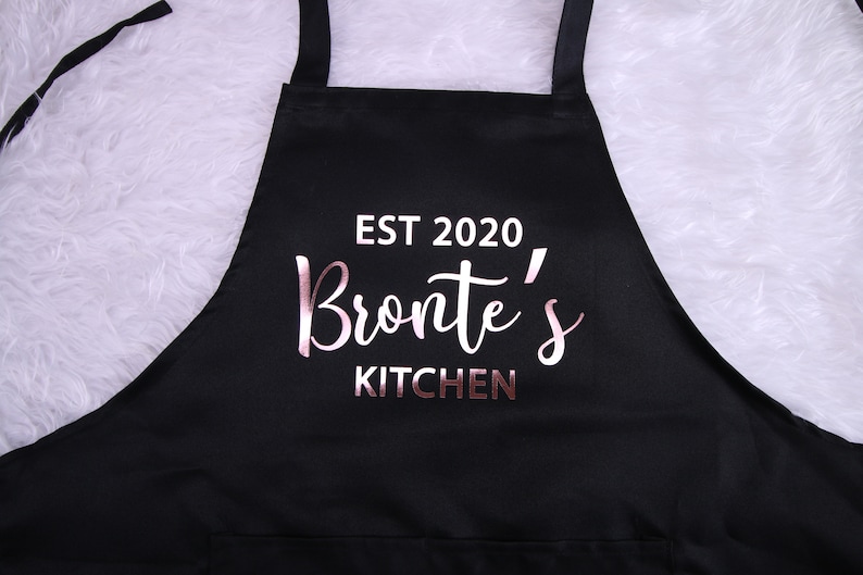 May include: Black apron with a white pocket and the text "EST 2020 Bronte's KITCHEN" in rose gold lettering.