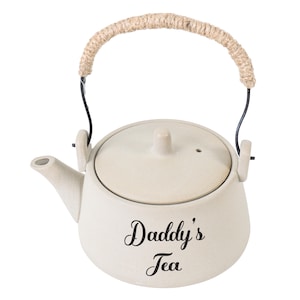Könnte beinhalten: Cremefarbene Keramik-Teekanne mit Deckel und Ausguss. Die Teekanne hat einen geflochtenen Griff und die Worte "Daddy's Tea" in schwarzer Schrift auf der Vorderseite. Eine charmante Ergänzung für jede Küche oder jedes Teeservice.