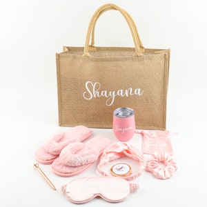 May include: A personalized gift set with a jute tote bag, pink fuzzy slippers, a pink tumbler, a pink satin sleep mask, a pink satin headband, a pink satin scrunchie, and a pink satin pouch. The tote bag, tumbler, slippers, sleep mask, headband, scrunchie, and pouch all have the name "Shayane" embroidered on them.
