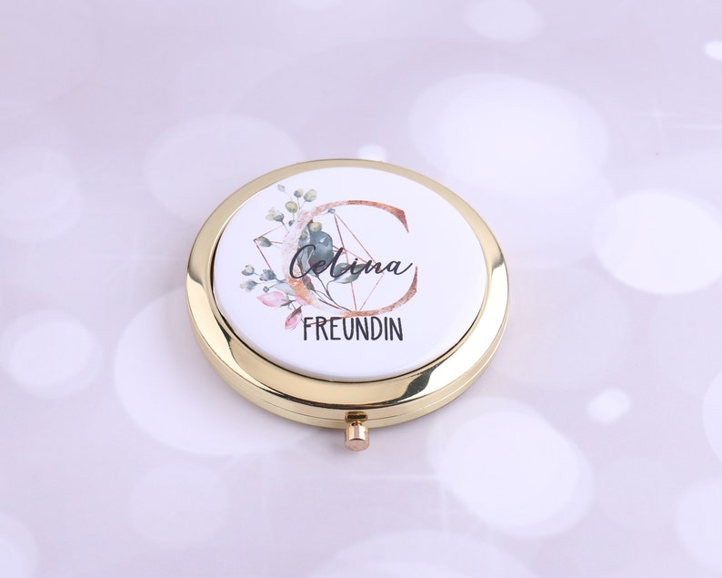 K&ouml;nnte beinhalten: Ein goldener Kompakt-Spiegel mit einem wei&szlig;en Deckel, der mit einem Blumendesign und dem Text "Celina Freundin" verziert ist.
