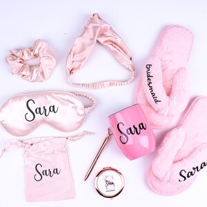 May include: A pink and gold bridesmaid gift set with the name "Sara" on a sleep mask, tumbler, and drawstring bag. The set also includes a pink headband, hair scrunchie, and fuzzy pink slippers with the word "bridesmaid" embroidered on one slipper.