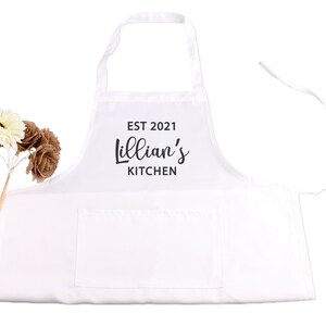 May include: White kitchen apron with a large pocket and adjustable straps. The apron has black text that reads "EST 2021 Lillian's Kitchen".