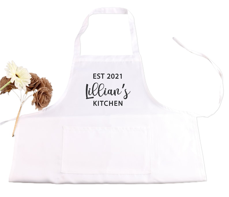 May include: White kitchen apron with a large pocket and adjustable straps. The apron has black text that reads "EST 2021 Lillian's Kitchen".