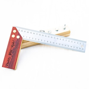 May include: A wooden try square with a reddish-brown finish and brass accents, featuring the engraved text 'Daddy, YOU Rule! Love, Arthur'. A stainless steel ruler with metric markings up to 20 cm is attached to the wooden handle with three screws.