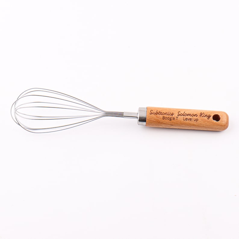 May include: A silver whisk with a wooden handle. The handle is engraved with the text "Subtronics Solomon King Boogie T Level Up".