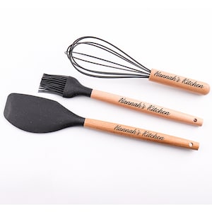 Peut inclure: Un ensemble de trois ustensiles de cuisine avec des manches en bois. Les manches sont gravés du texte "Hannah's Kitchen". Les ustensiles comprennent une spatule en silicone noire, un pinceau à badigeonner en silicone noir et un fouet en fil métallique noir.