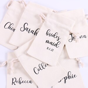May include: A set of eight white cotton drawstring bags with black lettering. The bags are labeled with names, including "bridesmaid", "Chia", "Sarah", "Jenna", "Rebecca", "Sophie", and "Collette". One bag also includes the date "11.1.22".