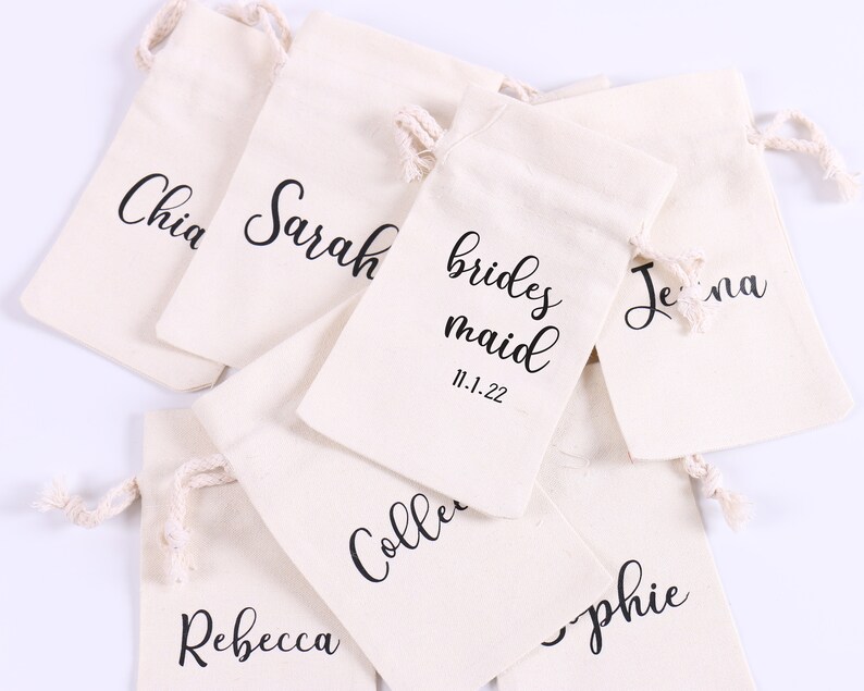 May include: A set of eight white cotton drawstring bags with black lettering. The bags are labeled with names, including "bridesmaid", "Chia", "Sarah", "Jenna", "Rebecca", "Sophie", and "Collette". One bag also includes the date "11.1.22".