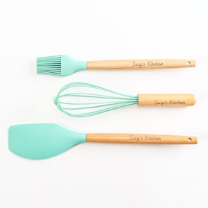 Może przedstawiać: Zestaw trzech zielonych narzędzi kuchennych z drewnianymi uchwytami. Uchwyty są grawerowane tekstem "Lucy's Kitchen". Zestaw zawiera silikonową szpatułkę, trzepaczkę i pędzel do smarowania.