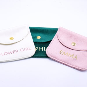Puede incluir: Tres pequeñas bolsas de tela con botones dorados y colores rosa, blanco y verde. Las bolsas tienen las palabras "Flower Girl", "Sophia" y "Emma" impresas en ellas.