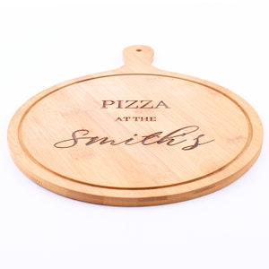 May include: A round, light brown bamboo pizza board with a handle. The board has a carved groove around the edge and the words "PIZZA AT THE Smith's" are engraved on the surface.