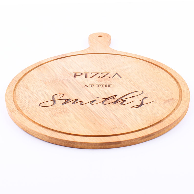 May include: A round, light brown bamboo pizza board with a handle. The board has a carved groove around the edge and the words "PIZZA AT THE Smith's" are engraved on the surface.