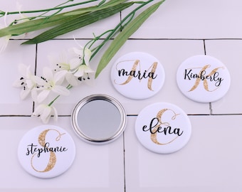 May include: Four round, white compact mirrors with gold glitter and black lettering. The names "Stephanie", "Maria", "Elena", and "Kimberly" are written on the mirrors.