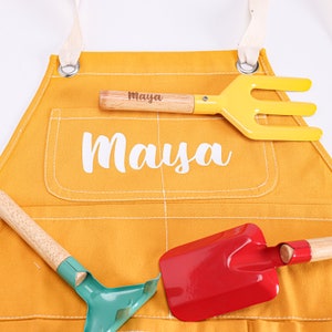 May include: A yellow gardening apron with a large pocket and the name "Maya" embroidered on it. The apron is shown with three gardening tools: a red shovel, a green rake, and a yellow hand rake.